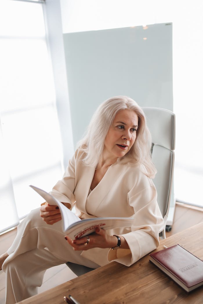 Elderly woman with white hair reading a book at a wooden desk in a bright office.